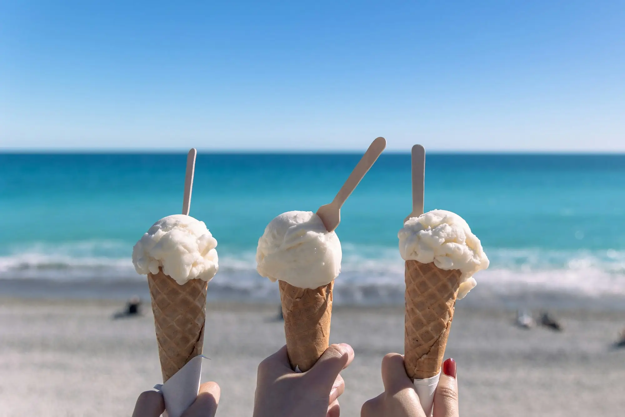 Three soft-serve ice creams held up against a sunny beach and ocean